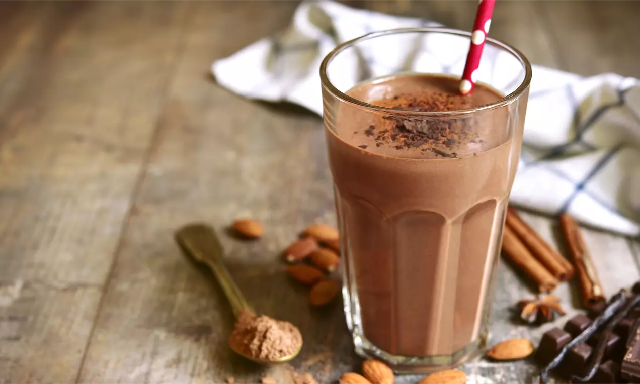 Frappè color cioccolato denso in bicchiere di vetro, decorato con cacao e cannella e circondato da mandorle e spezie su tavolo rustico
