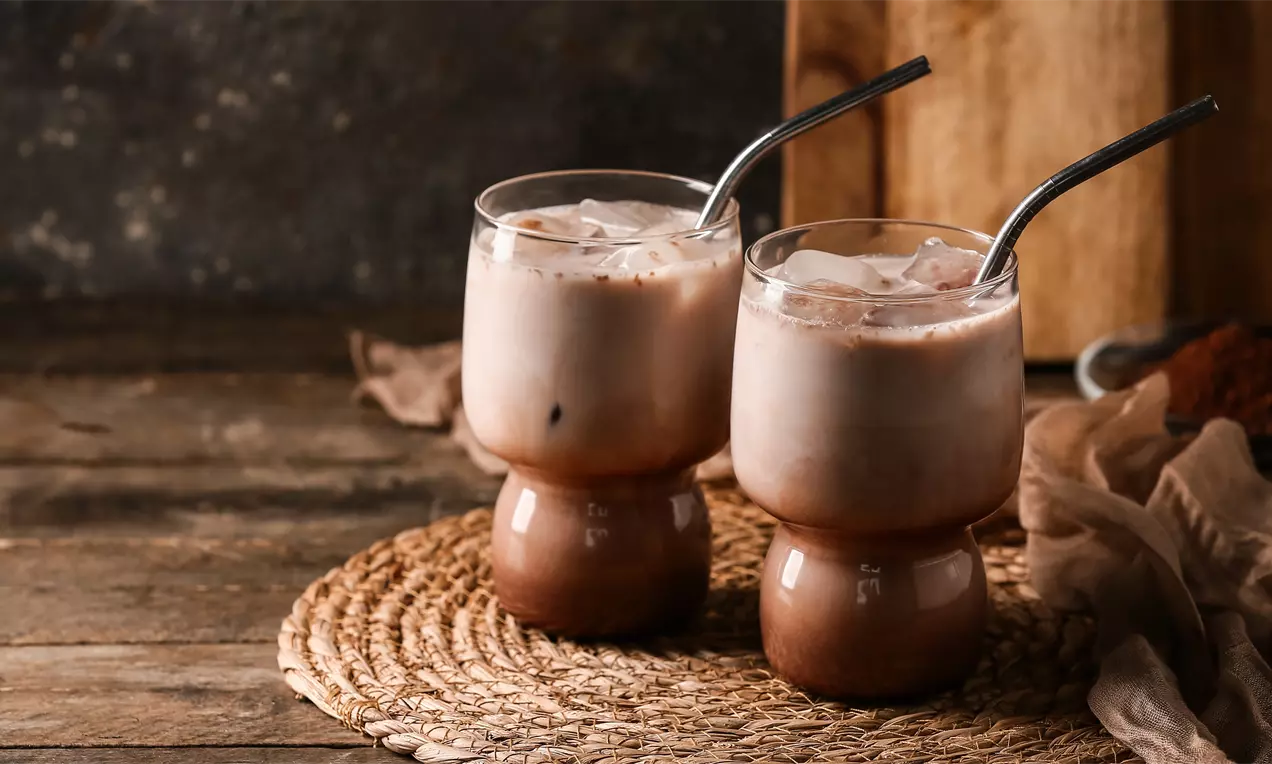 Due bicchieri di vetro su base arrotondata, colmi di shake cioccolato chiaro con ghiaccio, cannucce metalliche e fondo cacao su tappeto naturale