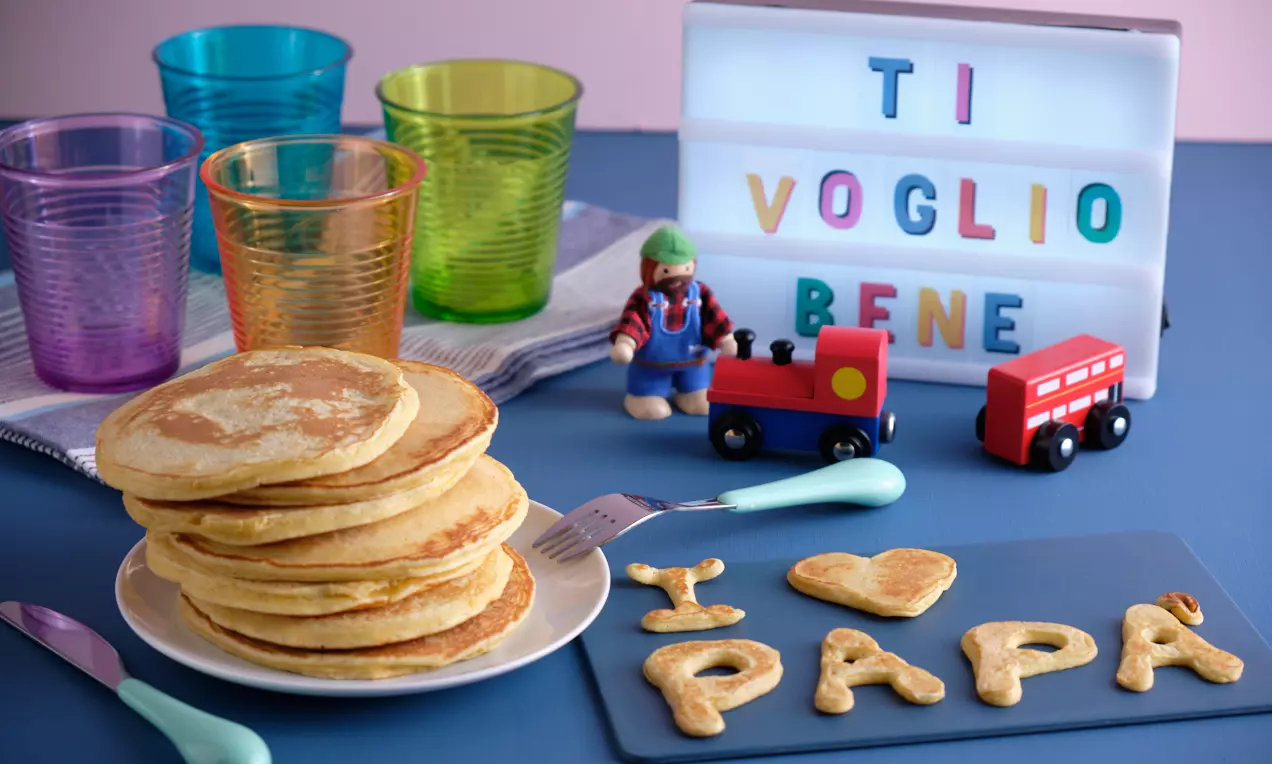 Pila di pancake dorati e soffici su un piatto bianco, accanto a scritte di pastella cotta “I ❤️ PAPÀ” su un vassoio nero