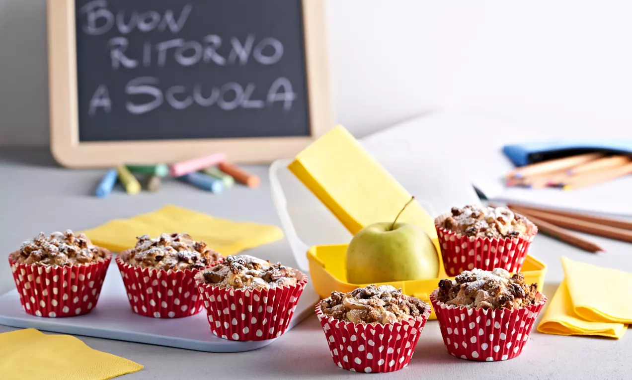 Muffin dorati con pezzetti di mela e crumble croccante di müesli, serviti in pirottini rossi a pois bianchi su un tavolo decorato con tovaglioli e una mela verde
