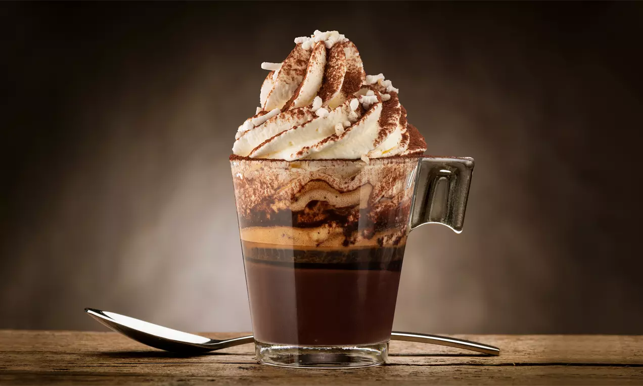 Un bicchiere trasparente con strati di cioccolato lucido, caffè cremoso e panna soffice decorata con cacao e scaglie bianche