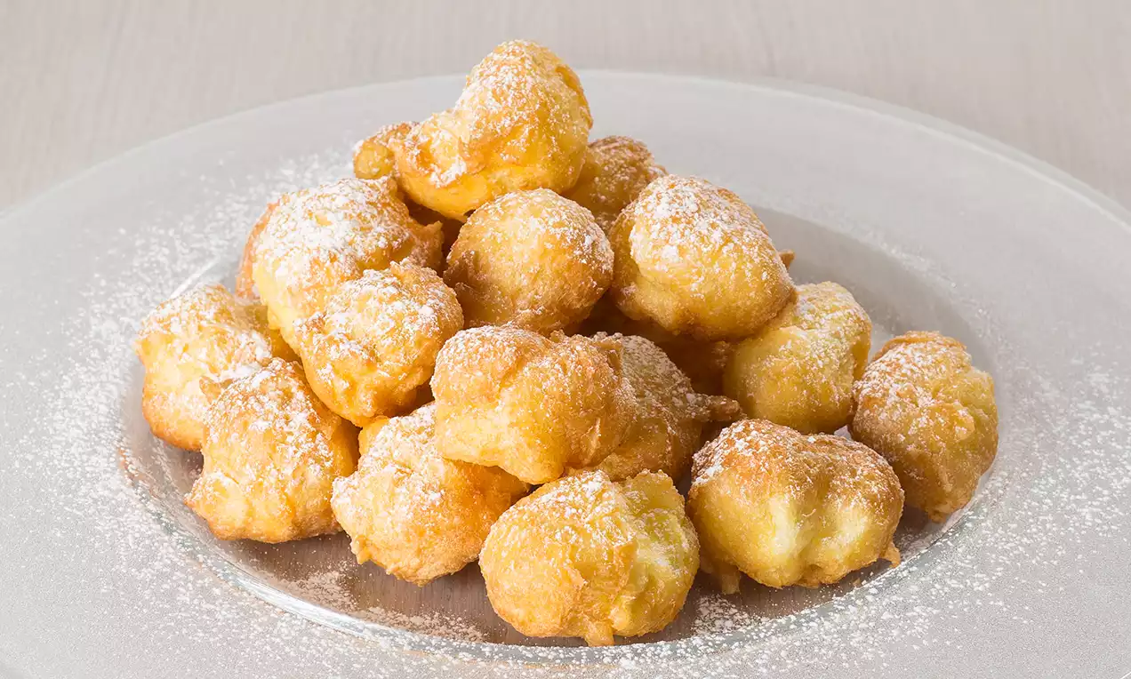 Frittelle dorate e gonfie, dalla forma irregolare, spolverate con zucchero a velo bianco, disposte a piramide su un piatto trasparente