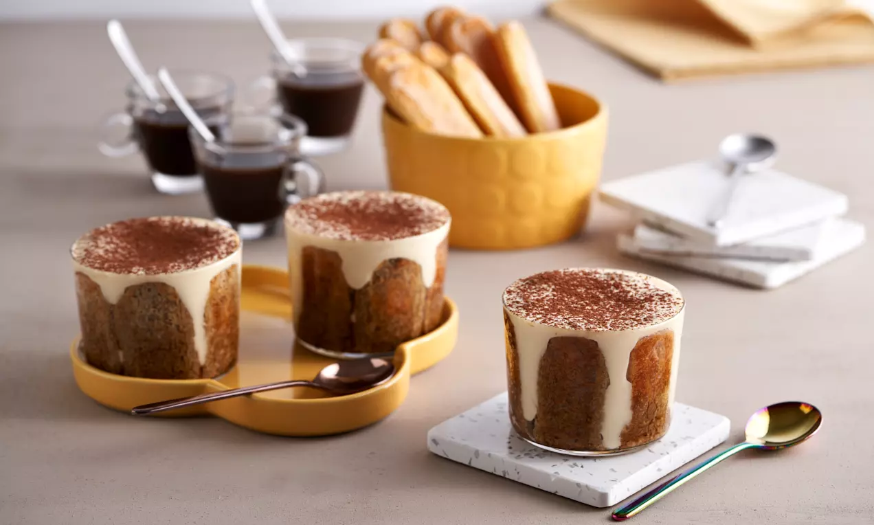 Bicchieri di tiramisù a strati, con crema morbida color avorio che cola sui bordi, spolverati di cacao e disposti su piatti color crema